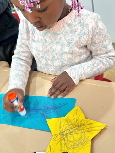 A child with colorful beads in their hair is seen gluing a blue piece of paper. In front of them is a yellow star-shaped paper with scribbles and a pen. The child appears focused on their craft activity at a table.