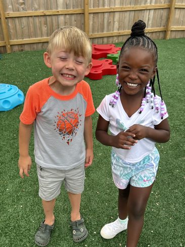 Two young children are smiling and standing on artificial grass near some colorful playground equipment, including a blue toy and red shapes. One child is wearing a gray and orange shirt with a basketball design, and the other is wearing a white shirt and patterned shorts.