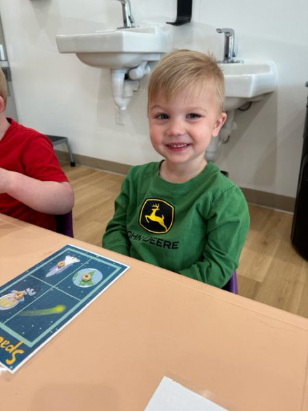 A young child with short blond hair sits at a table, smiling at the camera. The child is wearing a green shirt with a yellow deer logo and the text "JOHN DEERE." In front of the child is a card with space-themed illustrations. A sink is visible in the background.