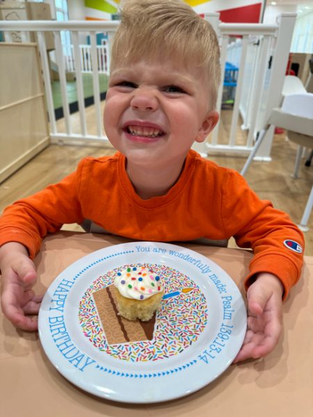 A young child with blonde hair and an orange shirt is smiling widely at the camera. They are seated at a table with a birthday-themed plate in front of them, which holds a cupcake decorated with white frosting and colorful sprinkles.