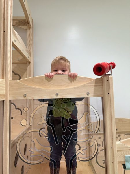 A young child, standing in a wooden play structure, peers over a panel featuring an engraved tree design. The child is grasping the top of the panel with both hands and appears to be smiling. The structure also has a red plastic toy telescope attached to it.