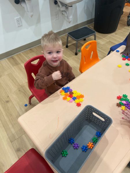 A young child with blond hair sits at a table with colorful interlocking flower-shaped pieces. The child is smiling and wearing a brown shirt, with an orange chair to the right and a red chair to the left. A gray basket is on the table, and wooden floors are visible.