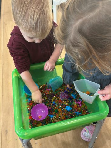 Two young children play with colorful water beads in a green sensory bin on a wooden floor. The child on the left uses a small blue scoop, while the child on the right uses a white scoop. Various other small tools and accessories are in the bin.