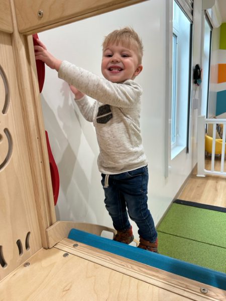 A young child with short blonde hair is smiling while playing on an indoor wooden climbing structure. They are wearing a light gray sweater, dark jeans, and brown shoes. The background features a colorful play area with bright green and yellow elements.