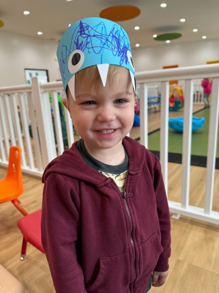 A cheerful young child wearing a blue hand-decorated paper crown with drawn-on eyes and teeth. The child is dressed in a maroon hoodie and is standing indoors in a brightly lit play area with colorful chairs and play structures visible in the background.