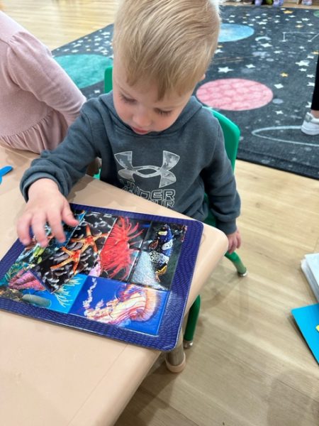 A young child with blonde hair is sitting at a small table, working on a colorful puzzle featuring sea creatures. The child is wearing a gray sweatshirt with the Under Armour logo. The background includes a colorful rug with a space-themed design and other children.