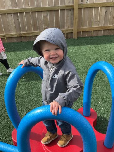 A young child wearing a gray hooded jacket smiles while standing on a red play structure. They are holding onto blue curved bars in an outdoor playground area with artificial grass and a wooden fence in the background.