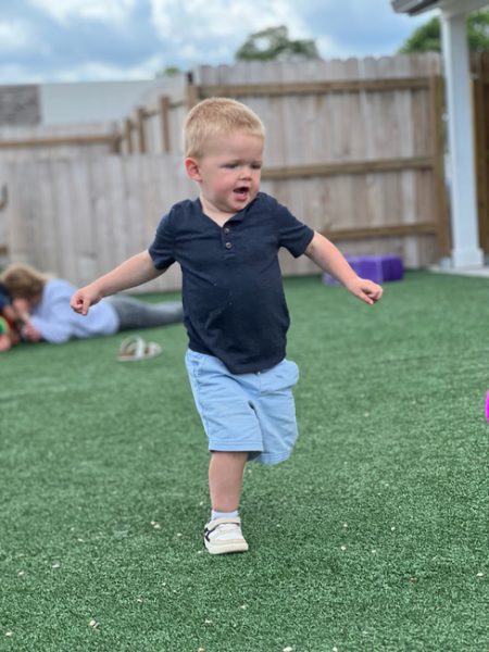 A young child with short blonde hair is running on a grassy lawn. He is wearing a navy blue shirt, light blue shorts, and white shoes. In the background, another child is lying on the grass near a wooden fence. The sky is partly cloudy.