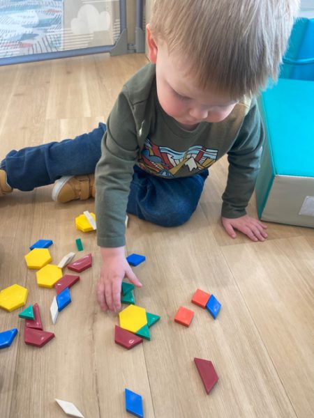 A young child in a long-sleeve shirt kneels on a light wooden floor while playing with colorful geometric shapes. The child is focused, using one hand to move the shapes. A blue cushioned play mat is partially visible on the right.