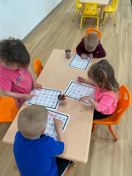 Four young children are sitting around a small table in a classroom, working on alphabet worksheets with crayons. The table and chairs are orange, and the floor is made of light-colored wood. The children appear focused on their activities.