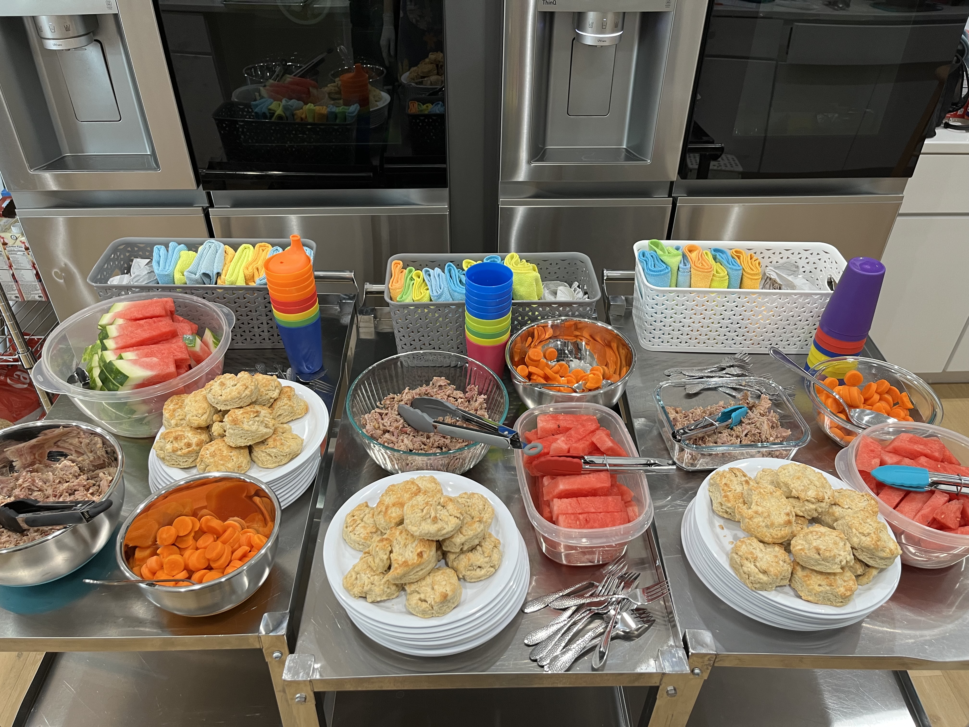 A buffet table with a variety of food items including watermelon slices, biscuits, carrot slices, tuna, and salads in bowls. Colorful cups are stacked next to the bowls, and napkins are rolled and placed in baskets behind the food. Spoons lie in front.