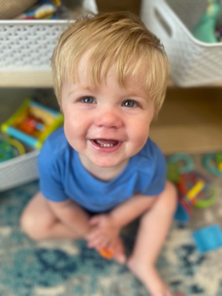 A young child with blonde hair sits on the floor in a blue shirt, smiling up at the camera. Behind the child are white woven baskets filled with toys. The child is surrounded by colorful play items.