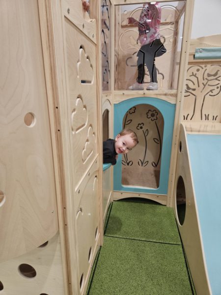 A young child peeks out of a small window in a wooden indoor playground structure with cloud and tree designs. Another child is partially visible on an upper level. The playground has green padded flooring and light blue accents.