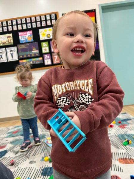 A young child with short hair smiles while holding a blue toy. The child is wearing a maroon sweatshirt with the word "Motorcycle" printed on it. In the background, another child is playing with red building blocks in a colorful room.