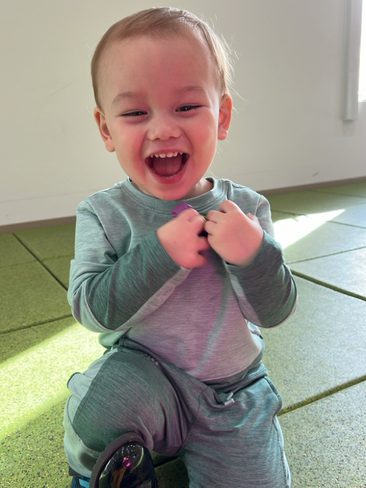 A joyful toddler with short hair, wearing a light green outfit and blue shoes, is kneeling on a green, tiled floor. The child is smiling widely and clutching the fabric of their shirt with small fists, illuminated by a soft light from a nearby window.