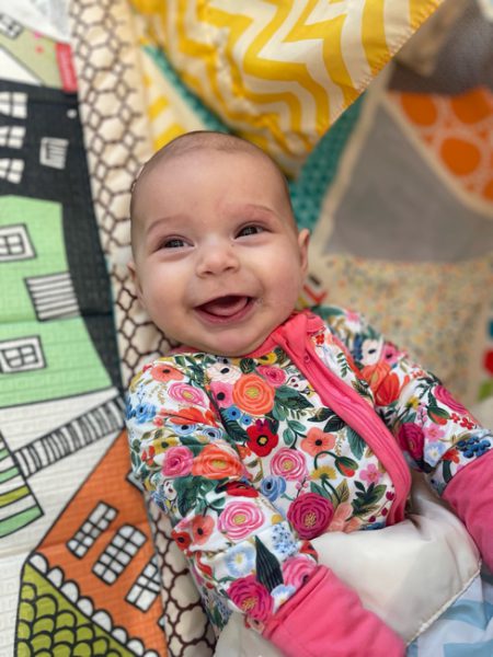 A happy baby with a big smile and wearing a colorful, floral outfit lies on a patterned quilt with house and geometric designs. The bright colors and playful background add to the cheerful mood of the image.