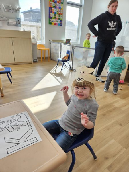 A young girl sits on a chair in a classroom, wearing a bunny mask she made from paper. A person in an Adidas hoodie stands behind her, supervising two other children who are playing. Tables and chairs are around the room, and sunlight streams through the windows.