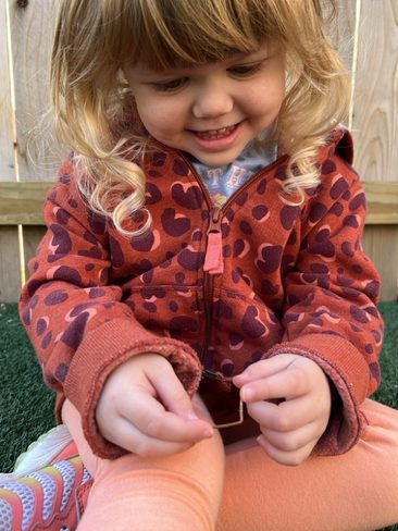 A young child with blonde curly hair and a smile wearing an orange hoodie with heart patterns and orange pants sits on a grassy surface playing with a small object. A wooden fence is in the background.