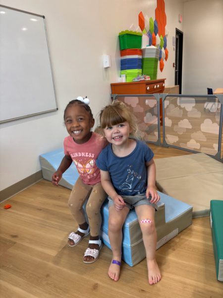Two young children are smiling while sitting on padded blocks in a brightly colored playroom. The child on the left wears a pink shirt and tights, while the child on the right wears a blue shirt and shorts. The room has a whiteboard and various play structures.