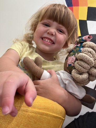 A smiling young child with light brown hair sits on a yellow chair, holding a doll with braided hair and a white dress. The background features a colorful, checkered wall pattern. The child's hand is reaching towards the camera.