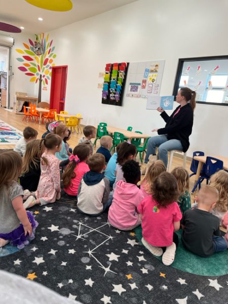 A teacher sits on a stool holding up a picture book in front of a group of young children seated on a colorful carpet in a classroom. The room is bright with colorful decorations, small tables, and chairs. A wall features a vibrant tree mural and hanging storage bins.