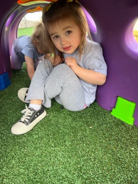 A young child with long blonde hair, dressed in a light gray shirt and gray pants, sits inside a colorful play tunnel on artificial grass. She looks directly at the camera with a curious expression. Another child, partially visible, is in the background within the tunnel.
