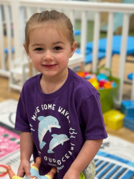 A young child with light brown hair is smiling while holding a toy. The child is wearing a purple shirt with images of dolphins and the text "Make Some Waves." In the background, there are toys and a play area with various colorful items.