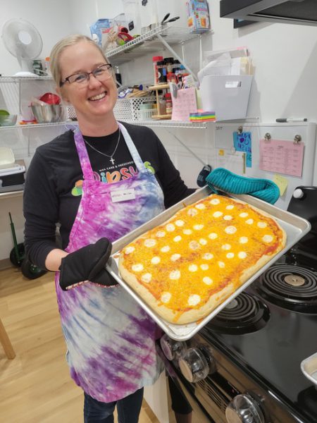 A woman wearing glasses and a colorful tie-dye apron smiles while holding a large rectangular pizza topped with cheese and pepperoni. She stands in a kitchen with a stovetop and various kitchen items and notes on the shelves and walls in the background.