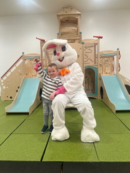 A young child stands beside a person in a white bunny costume, who is sitting. They are indoors, in front of a play structure with slides. The child is smiling and holding up a small object in their right hand. The floor is covered with green foam tiles.