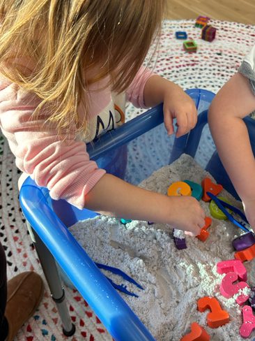 A young child with long hair plays in a blue sensory bin filled with sand and colorful plastic alphabet letters. The child is focused on the activity, wearing a pink long-sleeve shirt. Another child's arm is partially visible, also engaged in the play.