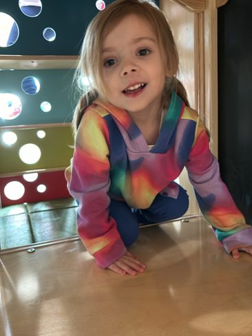 A young child with blonde hair smiles while crouching inside a colorful play structure. The child is wearing a rainbow-colored hoodie and blue pants. The play area features a wooden frame and panels with various circular cutouts.