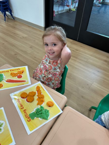 A young child with blonde hair and a floral dress sits at a table, smiling at the camera. On the table, there are playdough shapes and a placemat with colorful pictures of carrots. Behind them, other tables and chairs are visible against a background of wooden flooring.