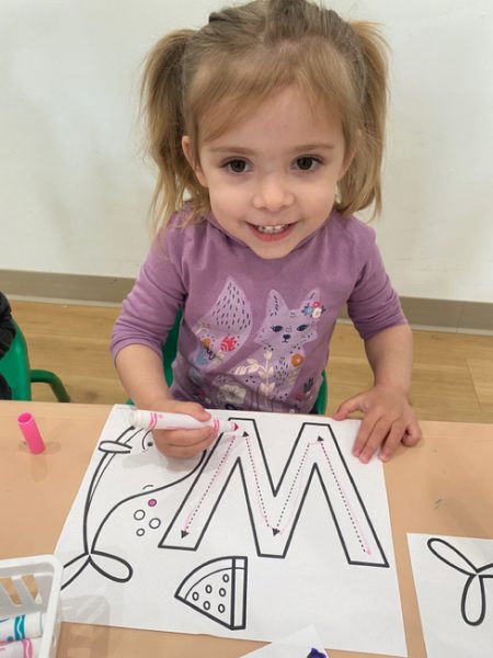 A young girl with light brown hair, wearing a purple shirt with animal prints, is sitting at a table and smiling. She is coloring a worksheet with the letter "M" on it using a pink marker. There are other craft supplies on the table.