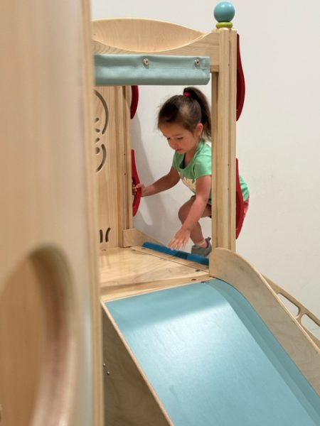 A young child in a green shirt and pink shorts is playing on a wooden indoor playground set. She is climbing a small structure with a blue ramp, holding onto red handlebars as she navigates her way. The background is plain and white.