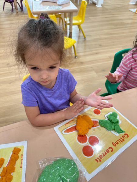 A young child wearing a purple shirt is sitting at a table, playing with colorful playdough. In front of her is a placemat with words "Cutting Carrots" and various carrot-related shapes. She is rolling orange playdough between her hands. Other children are visible in the background.