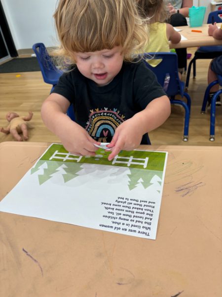 A toddler with curly blonde hair is standing at a table, joyfully working on a craft project involving a paper with a green forest scene and a nursery rhyme. Other children and a baby doll are in the background. The toddler is focused and concentrating.