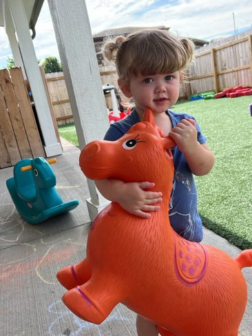 A young child stands outdoors on a concrete patio holding an orange inflatable toy with a horse design. The child has light brown hair styled in pigtails and is wearing a blue outfit. Chalk drawings are visible on the ground, and play equipment is in the background.
