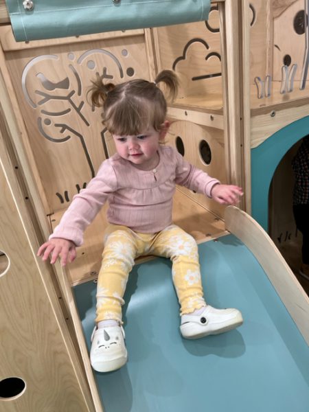 A young child with pigtails, wearing a pink long-sleeve shirt, yellow floral pants, and white shoes, sits at the top of a play slide. The play area is made of light-colored wood with carved designs of trees and clouds. The child appears ready to slide down.