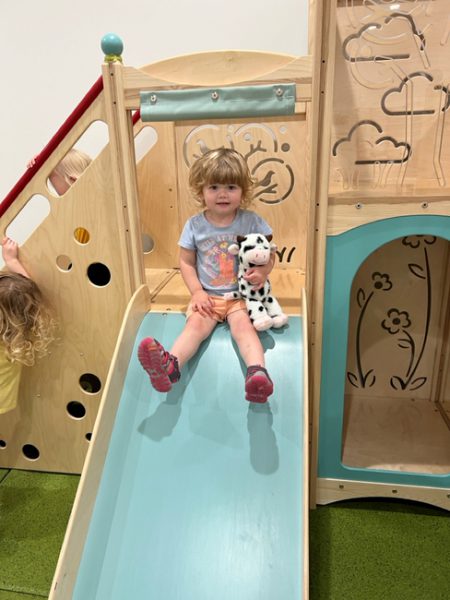 A young child with short blonde hair sits at the top of a blue slide holding a black and white spotted stuffed cow. They are wearing a light blue T-shirt with graphics and pink shoes. Another child is partially visible to the left, also playing. The background shows a wooden play structure.