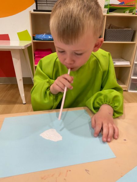 A young child in a green smock is sitting at a table, using a white straw to blow on a white substance placed on a light blue paper. The background shows some colorful shelves and various art supplies.