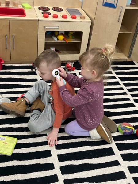 Two children playing on a black-and-white striped rug. One child, wearing a purple sweater and light purple pants, is holding a toy thermometer to the ear of another child who is sitting in front, wearing a red long-sleeve shirt and gray pants. A play kitchen is in the background.