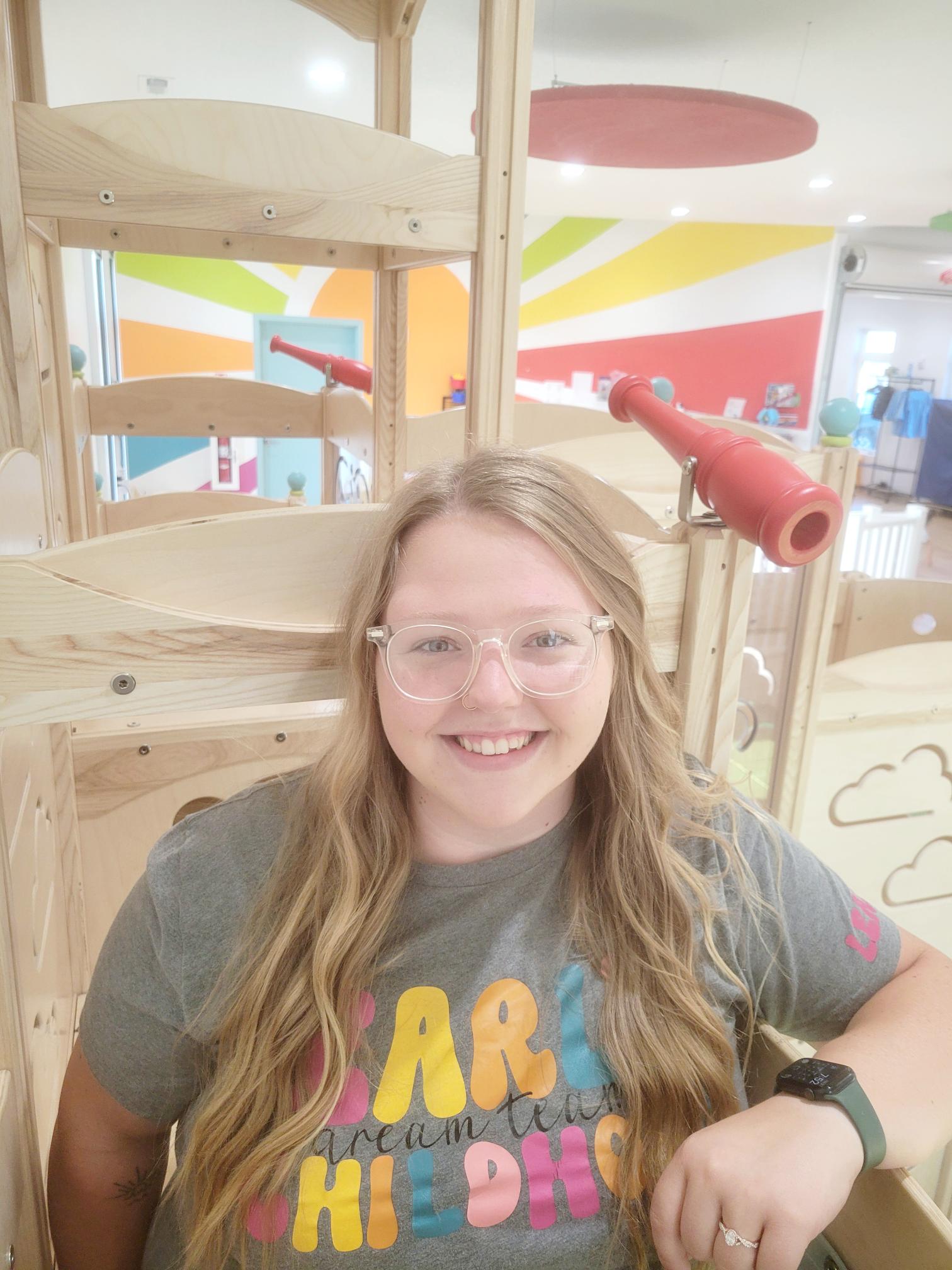 Sam in a grey t-shirt and clear glasses sitting on a large wooden jungle gym