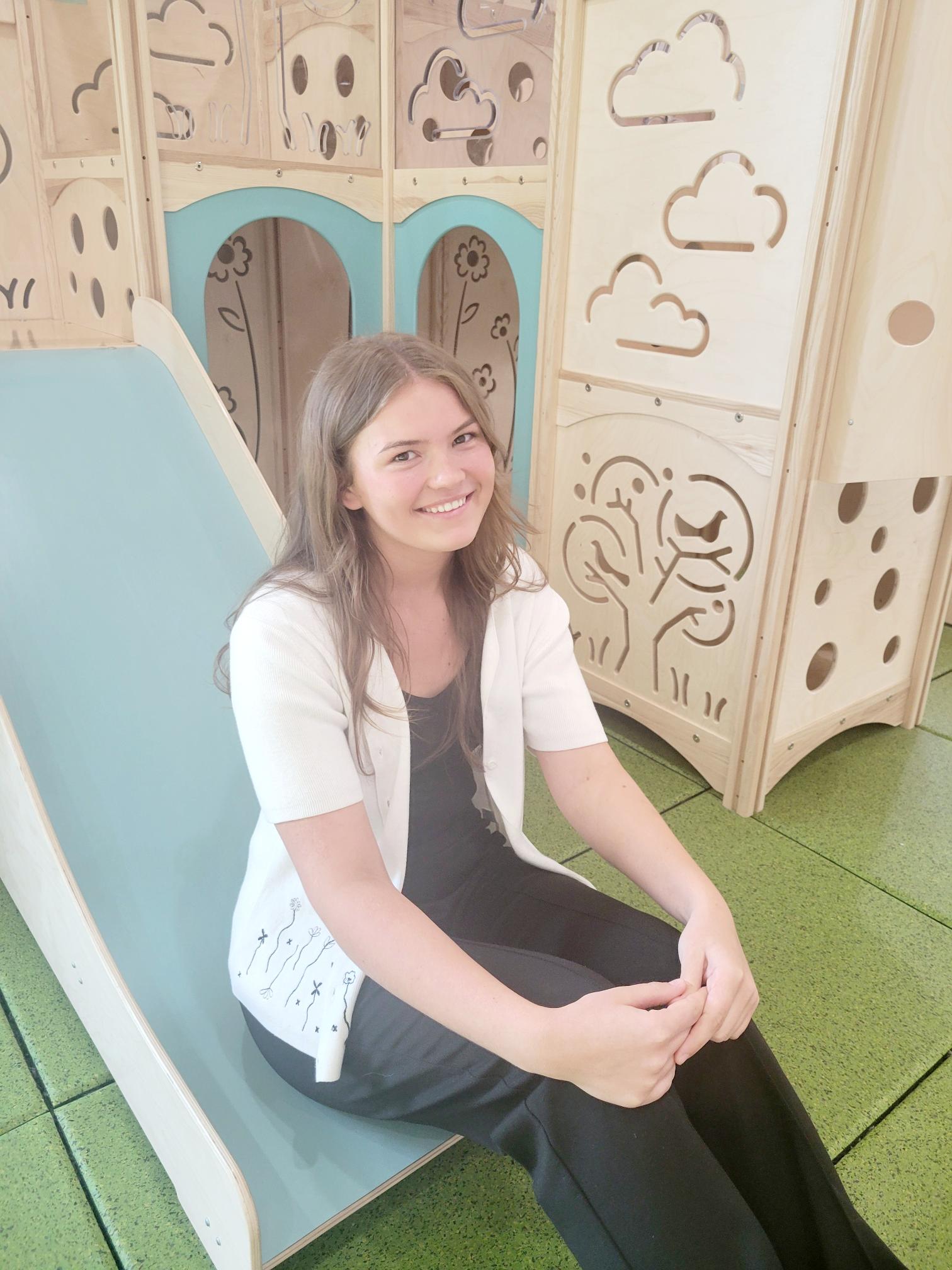 Anna wearing a white sweater over a black tank top and black pants sitting on the slide of a large wooden jungle gym