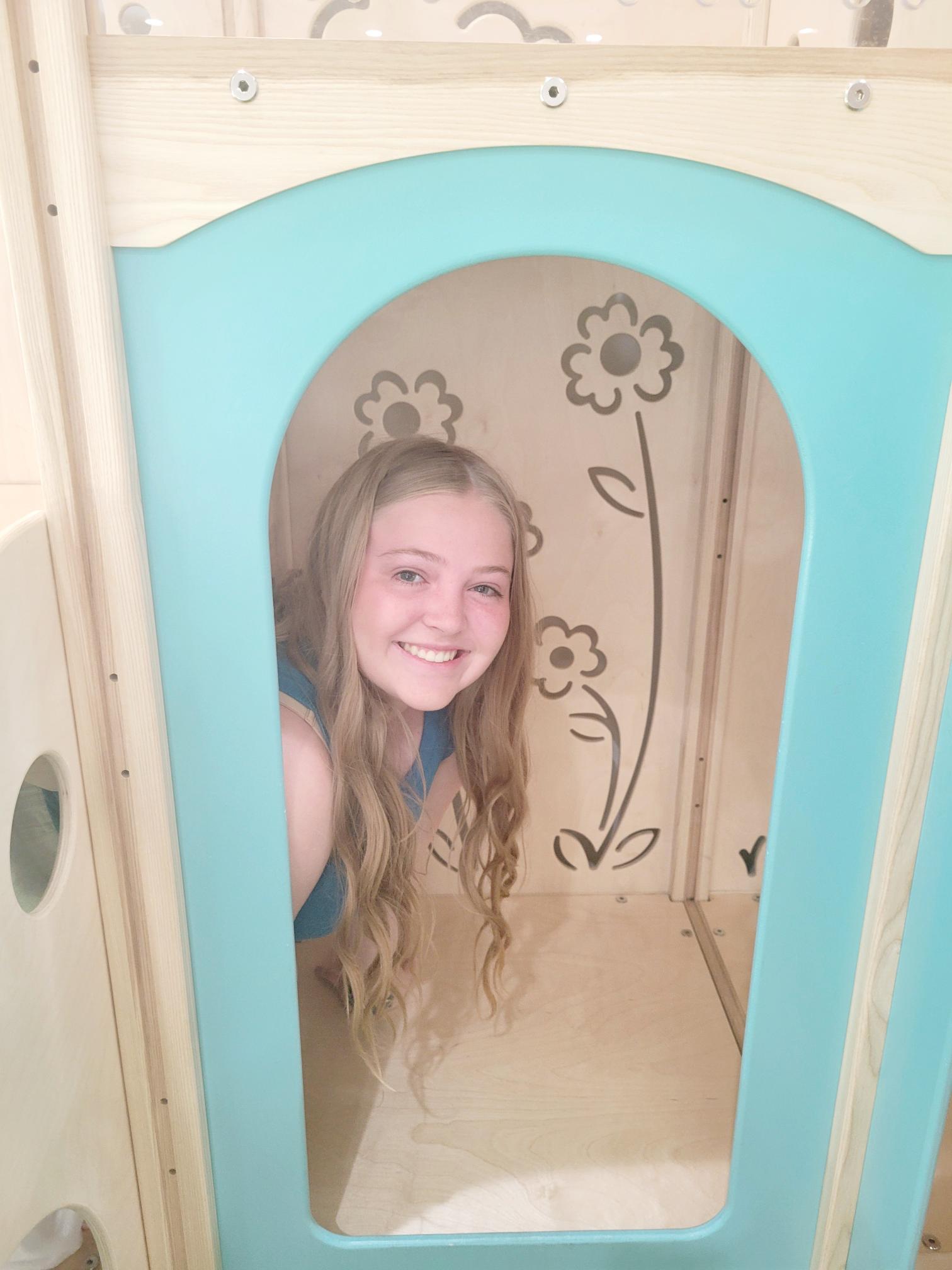 Peyton wearing a blue shirt smiling while inside a small wooden playhouse. The interior walls are decorated with simple flower drawings, and the playhouse has a light blue arch-shaped doorway