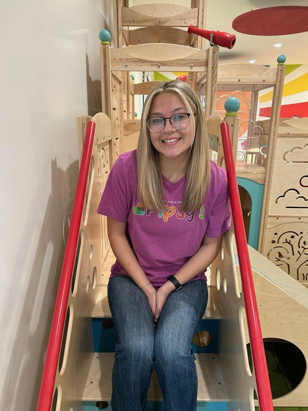 A person with long hair and glasses is sitting on a colorful indoor playground structure. They are wearing a purple t-shirt and jeans, smiling at the camera. The background features playful designs in a bright and cheerful setting.