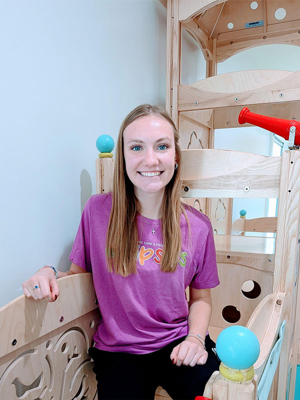 Woman smiling and sitting on a wooden playground structure indoors, wearing a purple T-shirt and black pants. The play area includes colorful balls and tubes, and she appears relaxed and happy.