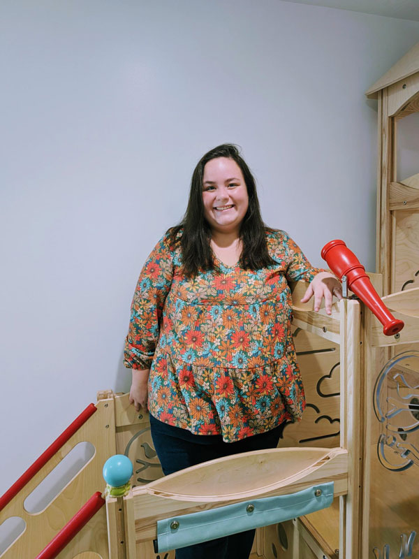 A woman with long dark hair stands inside a children's play structure with wooden elements. She is smiling and wearing a colorful floral blouse and dark pants. A red telescope is mounted nearby. The background is plain and bright.