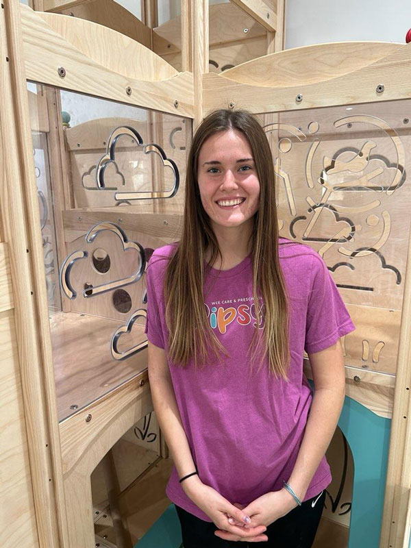 A woman with long brown hair wearing a pink t-shirt stands smiling in front of a wooden play structure. The structure has transparent panels with cloud and swirl designs.