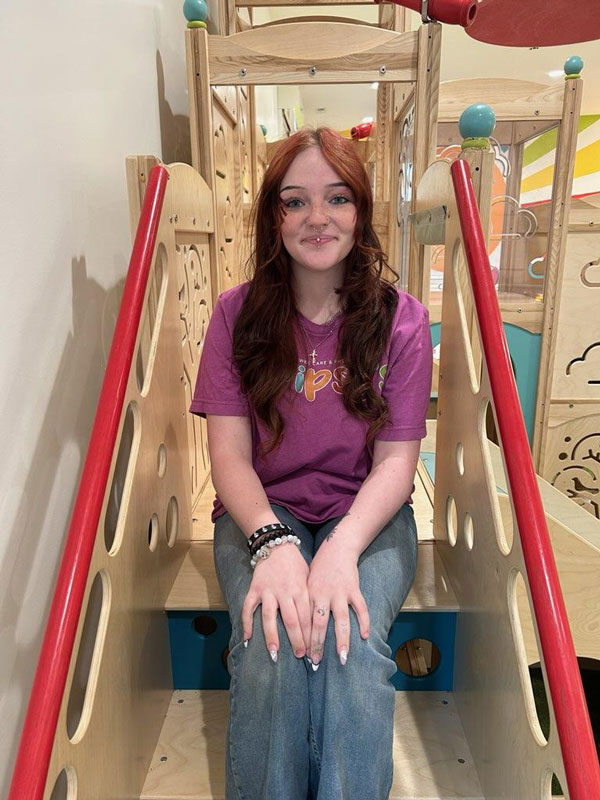 A person with long red hair sits on a wooden play structure indoors, wearing a purple shirt and jeans, smiling at the camera. The background features colorful decorations and playful elements.