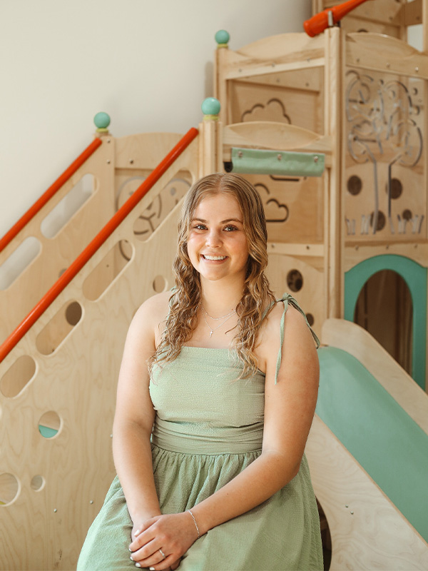 A young woman with wavy blonde hair, wearing a green sleeveless dress, sits and smiles in front of a wooden indoor playground structure with slides and climbing features.